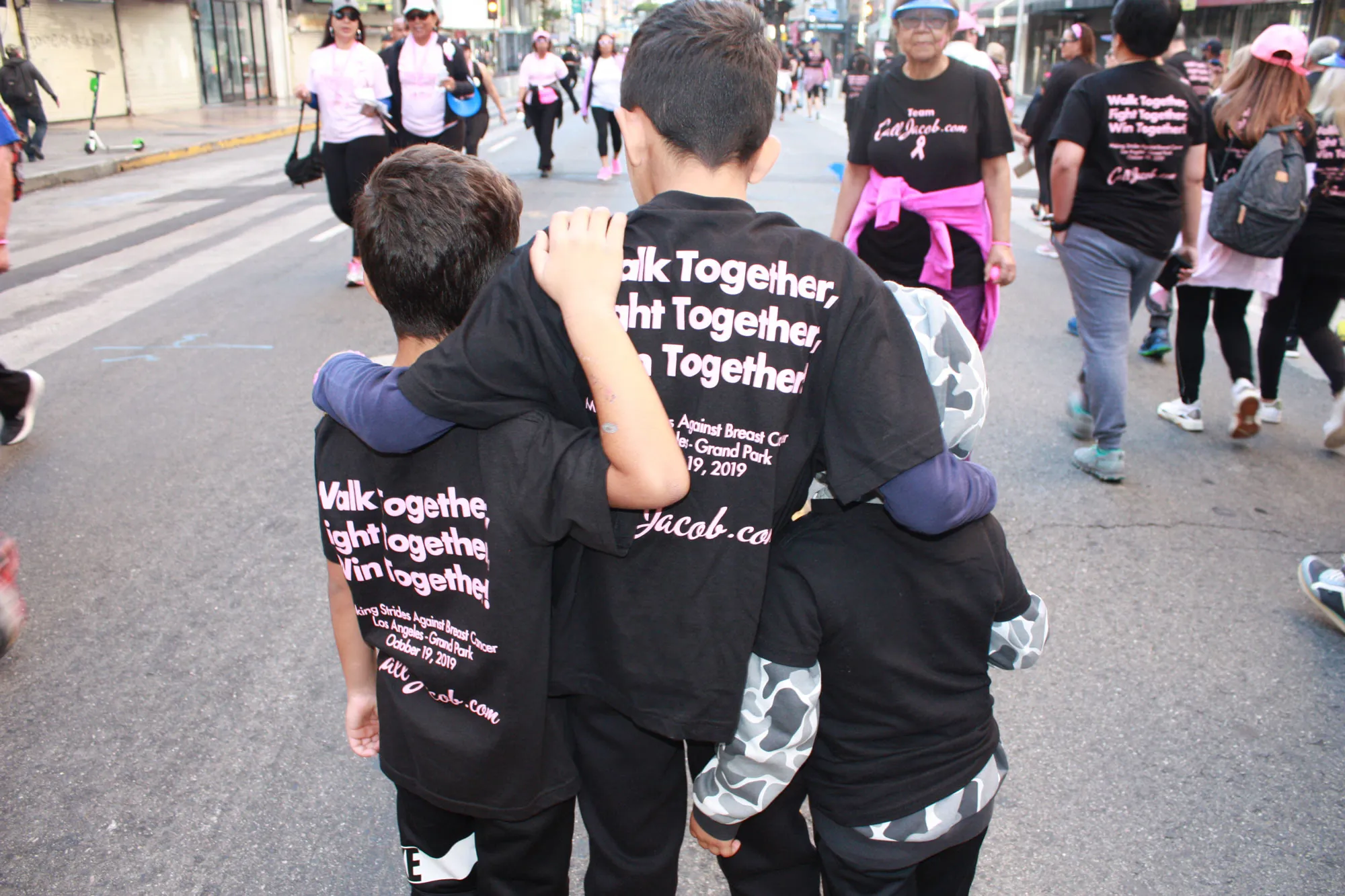 Three children with their arms around each other walk down a city street during an event. They wear matching black shirts reading, "Walk Together, Fight Together," as part of a group supporting personal injury awareness.