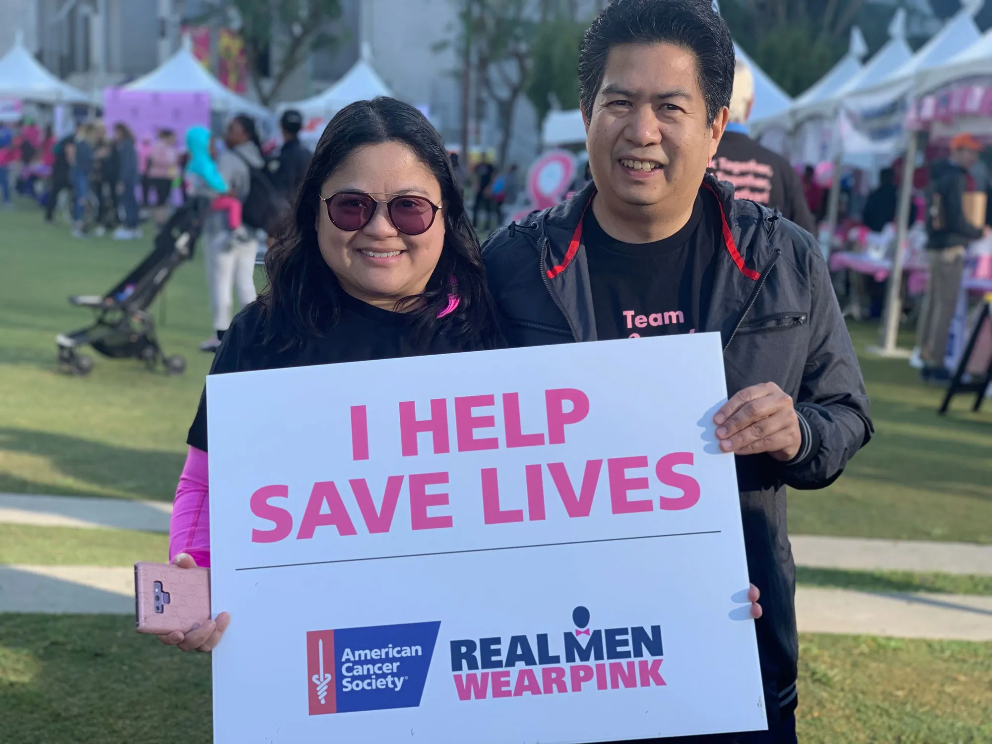 Two people smiling and holding a sign that says "I HELP SAVE LIVES" at an outdoor event to raise awareness about car accidents. The sign features the American Cancer Society and Real Men Wear Pink logos. Tents and participants are visible in the background.