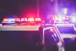 A row of police cars with flashing red and blue lights illuminates the night, creating a blurred, dramatic scene. The focus is on the emergency lights atop the vehicles.