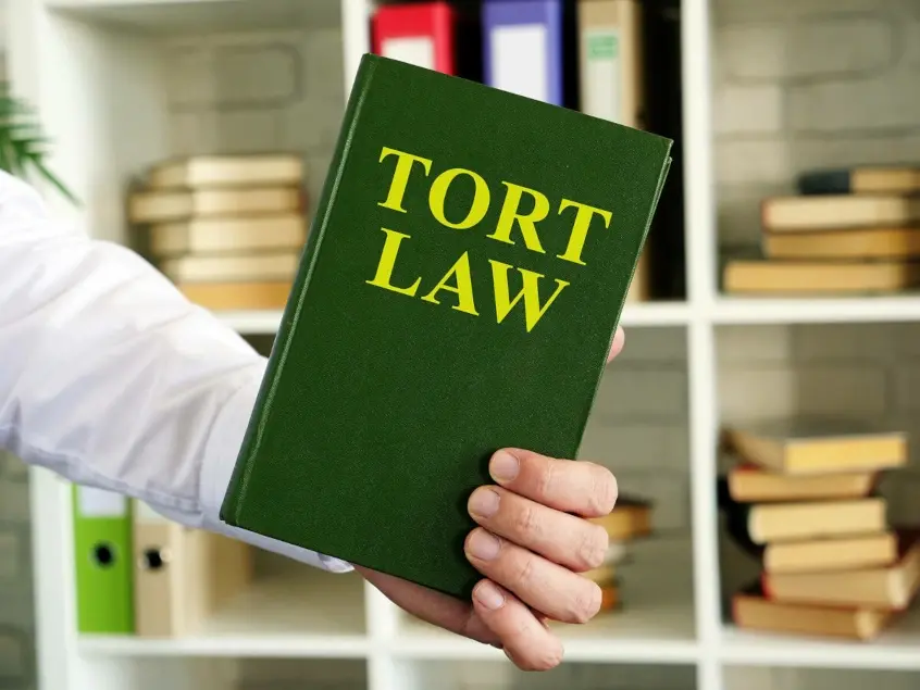 A hand holding a green hardcover book with the title "TORT LAW" in large yellow letters, in front of a bookshelf filled with books.