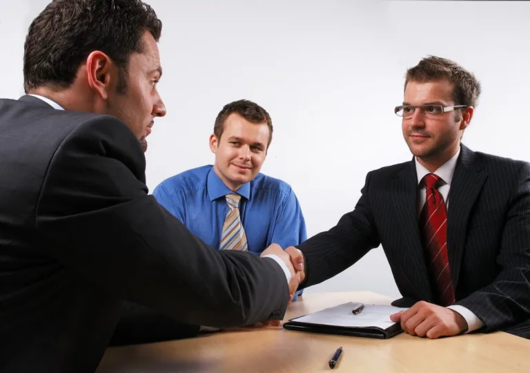 Three men in business attire sit at a table; two are shaking hands while the third man smiles, suggesting a successful meeting with a personal injury attorney. A pen and document on the table hint at resolving a car accident case.