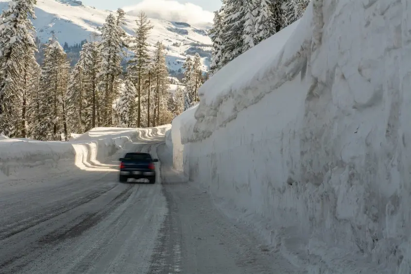 A car drives on a snow-covered mountain road flanked by tall snow banks, with snow-laden trees and mountains visible in the background.