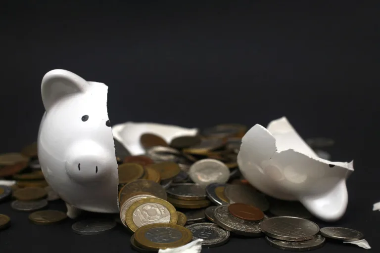 A broken white piggy bank is surrounded by scattered coins on a dark background, its shattered pieces echoing the unexpected loss often faced after car accidents.
