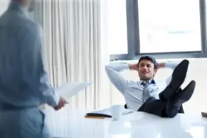 A man in a suit sits at a desk with his feet up and hands behind his head, looking relaxed. Another person stands nearby holding documents. The setting appears to be a modern office.