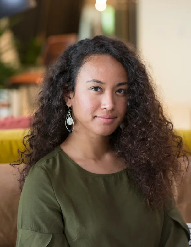 A woman with long curly hair, wearing a green top and teardrop earrings, sits indoors on a couch, looking at the camera with a slight smile. The background is softly blurred.