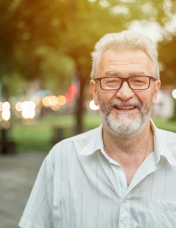An older man with gray hair, a beard, and glasses smiles while standing outdoors in a park. The background is softly blurred with green trees and bokeh lights. He wears a light striped shirt.