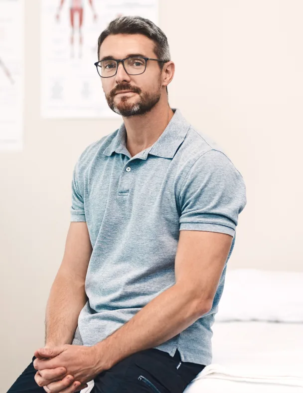 A man with short dark hair, a beard, and glasses sits on the edge of an examination table, wearing a light gray polo shirt. The background shows blurred anatomical posters on a cream-colored wall.
