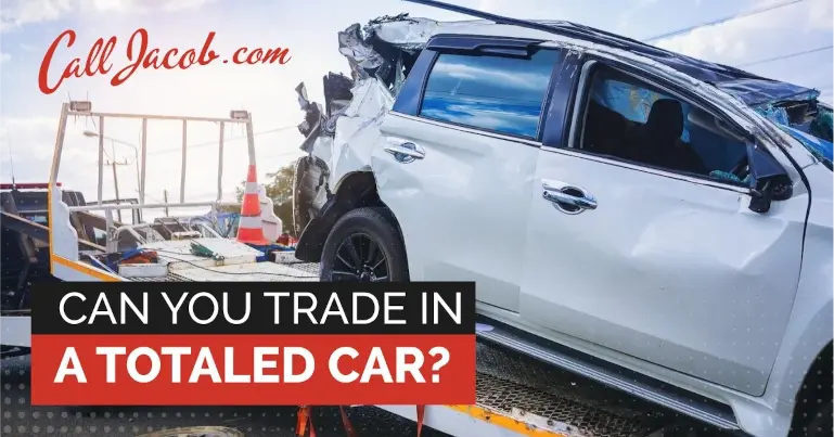 A severely damaged white SUV is loaded onto a tow truck. Text reads: "Call Jacob.com" and "Can you trade in a totaled car?" under bold red and black banners.