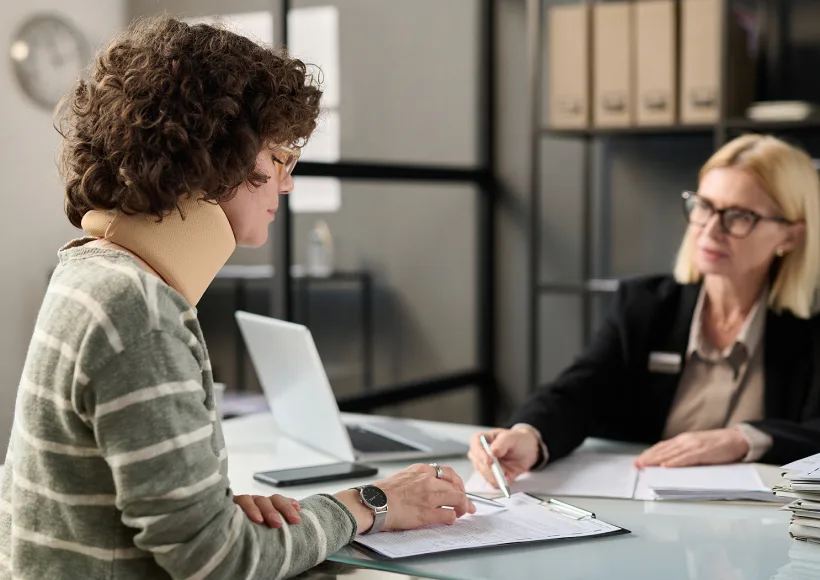 A woman in a neck brace sits at a desk, filling out paperwork with the help of a car accident attorney in business attire, who is pointing at a document. Both are in an office setting.