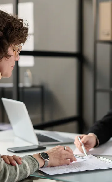 Two people sit at a desk with papers, one pointing at a document with a pen. A laptop is open on the table, suggesting a meeting with lawyers or a car accident attorney in an office or meeting room.