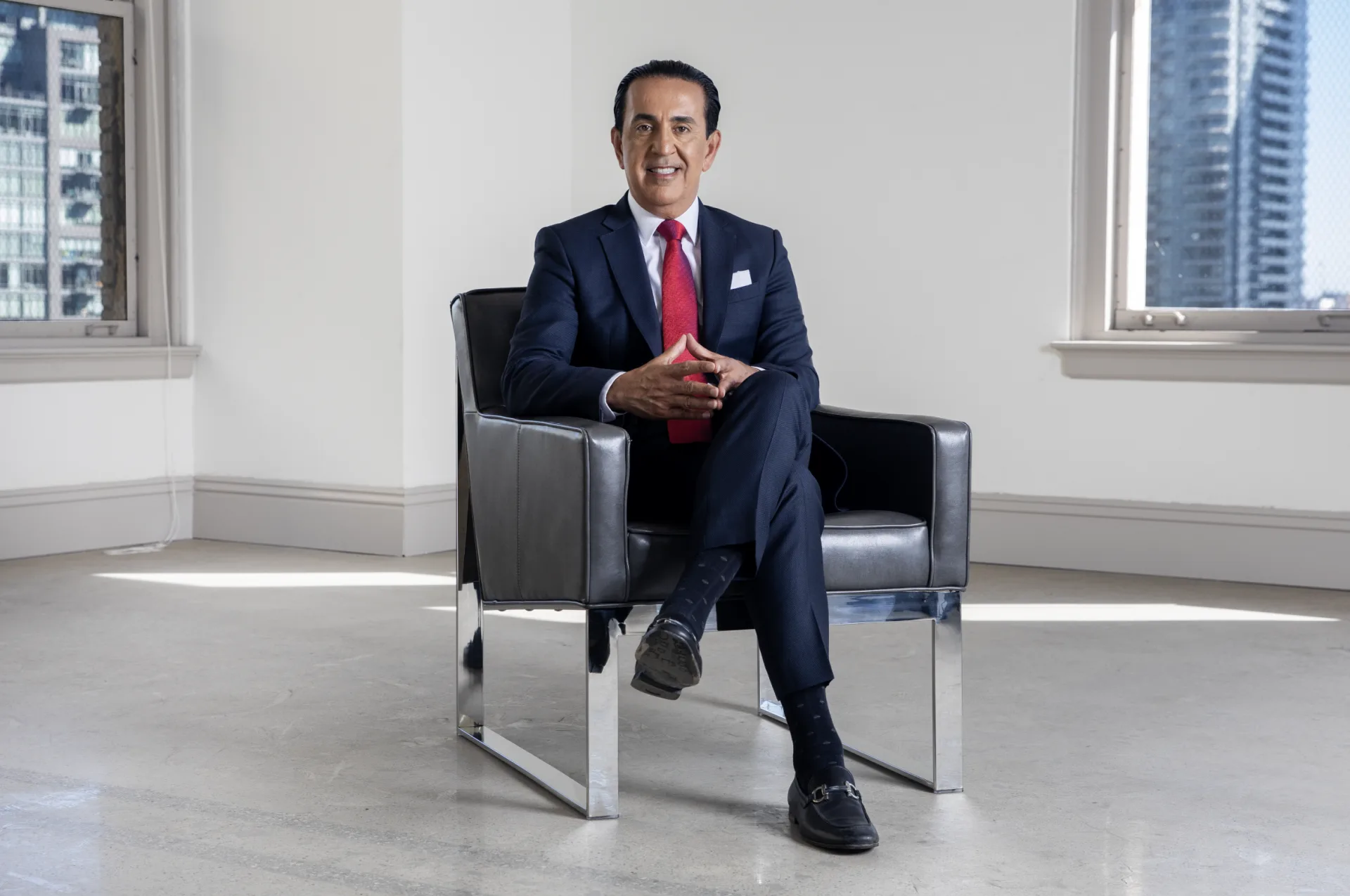 A personal injury attorney in a dark suit, red tie, and black shoes sits confidently in a modern black armchair in a bright city office with large windows. He is smiling, with hands clasped and legs crossed.