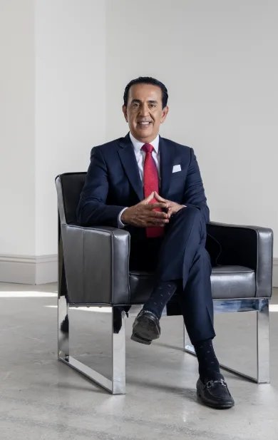 A man in a dark suit with a red tie, perhaps a personal injury attorney, sits in a modern metallic armchair, smiling with his hands clasped. The setting is a bright room with light walls and a concrete floor.
