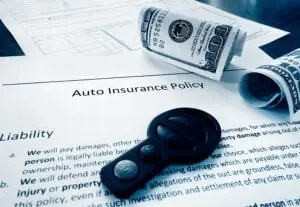 A car key, rolled-up cash, and paperwork labeled "Auto Insurance Policy" are shown on a table, symbolizing car insurance and financial planning.