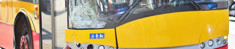 A yellow city bus with a shattered front windshield is parked near a red surface. The cracked glass on the driver's side hints at an accident—personal injury attorneys may be needed if anyone was hurt. Accessibility stickers are visible by the door.