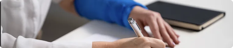 A person wearing a blue sleeve writes on paper with a pen at a desk, possibly jotting down details about car accidents, while a closed black notebook lies nearby.