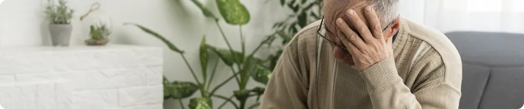 An elderly man sits indoors on a couch, wearing a beige sweater and glasses, resting his head in one hand, appearing stressed or deep in thought—perhaps after dealing with personal injury from car accidents. Green plants and white decor fill the background.