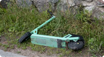 A damaged green electric scooter lies on its side in the grass near a stone wall, with the handlebar and wheels visible—an image that highlights why consulting a personal injury or car accident attorney can be crucial after such incidents.