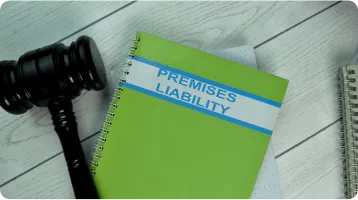 A green spiral notebook labeled "Premises Liability" lies on a white wooden surface next to a black judge's gavel and another partially visible notebook, highlighting the importance of personal injury knowledge for any personal injury attorney.