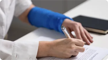 A person with a blue arm cast is sitting at a desk, filling out a form with a pen—perhaps seeking help from personal injury attorneys. A closed notebook rests nearby on the white surface.