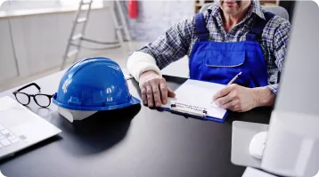 A person in blue overalls sits at a desk, writing on a clipboard. A blue hard hat and glasses are on the desk nearby, with office items in the background—suggesting careful documentation, much like a personal injury attorney preparing a case.