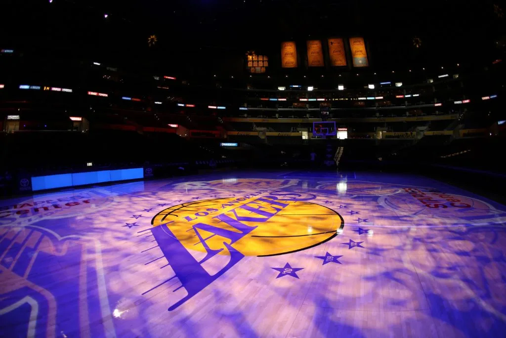 An empty basketball court with a large Los Angeles Lakers logo illuminated at center court, surrounded by empty seating and championship banners above—silent, as if awaiting a verdict from top lawyers or a car accident attorney.