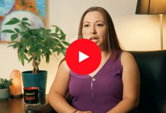 A woman in a purple sleeveless top sits in an armchair, speaking. Behind her are a plant, lamp, framed artwork, and a mug on a table—creating the perfect setting for an informative personal injury attorney video. A red play button overlays the center.