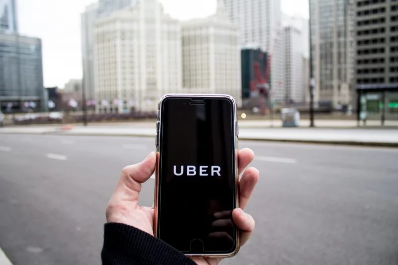 A person holds a smartphone displaying the Uber app logo on the screen, with tall city buildings and an empty street in the background—an important scene for lawyers handling car accidents in urban areas.