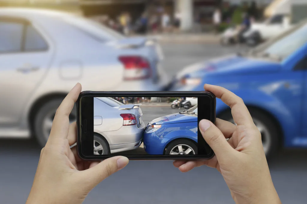 A person holds a smartphone to take a photo of a car accident, capturing a silver car and a blue car that have collided bumper-to-bumper on a street—evidence often needed by a car accident attorney.