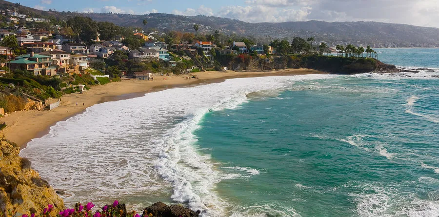 A scenic coastal view of a sandy beach bordered by turquoise waves and rocky cliffs, with houses and greenery on the hillsides under a partly cloudy sky—perfect for relaxing or meeting with personal injury attorneys in a serene setting.
