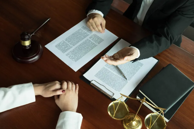 Two people in business attire sit across a wooden desk with legal documents, a gavel, scales of justice, and a notebook—one gestures while the other listens with hands folded, suggesting a car accident attorney consultation.