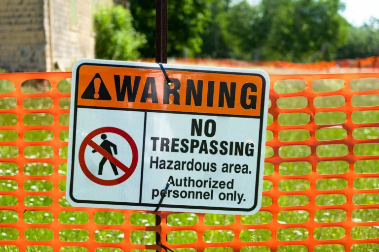 A “Warning” sign on orange mesh fencing reads: “No Trespassing. Hazardous area. Authorized personnel only,” with a symbol of a person barred from entry. Such areas often require a personal injury attorney if accidents occur. Green grass and trees are visible in the background.