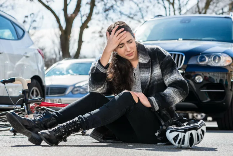 A woman sits on the ground in a parking lot, looking pained and holding her head. A bicycle and helmet are nearby, suggesting a fall. Personal injury from car accidents or bike incidents can happen anytime—always seek help if you’re hurt.