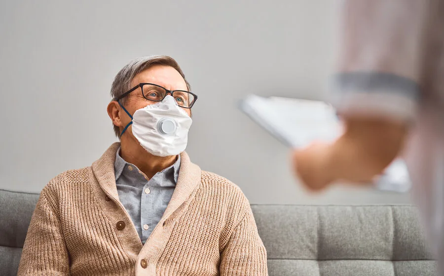 An older man wearing glasses and a mask with a valve sits on a gray couch, looking at a person holding a clipboard—possibly consulting with personal injury lawyers—in the foreground.