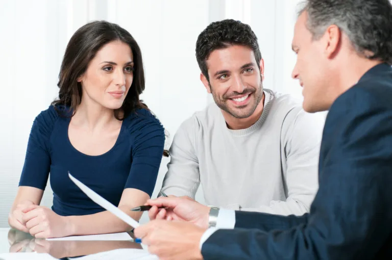 A woman and a man sit together at a table, smiling and listening to another man in a suit holding documents and a pen, suggesting a professional meeting with lawyers about personal injury or car accidents.