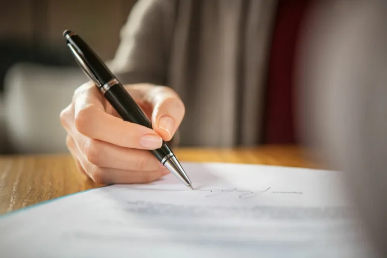 Close-up of a person's hand holding a black pen and signing a document on a wooden table—ideal imagery for lawyers handling cases like car accidents. The focus is on the pen and signature, with the background softly blurred.