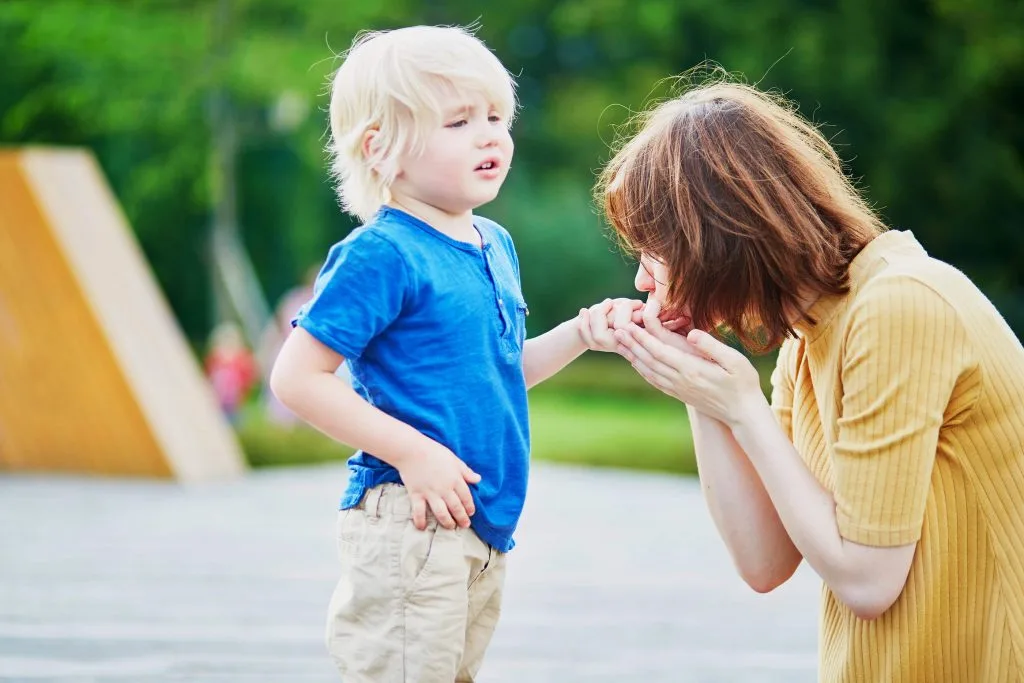 A woman kneels and kisses a young child’s hand outdoors. The child, in a blue shirt and beige shorts, looks upset—perhaps after a minor fall. Even small accidents like these remind us how personal injury attorneys support families.