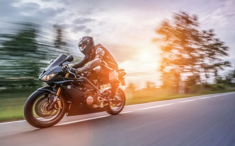 A motorcyclist in black gear and helmet rides a black sport bike at high speed on a road, with blurred trees and sunlight in the background, conveying motion and speed—reminding us how crucial car accident attorneys can be after accidents.