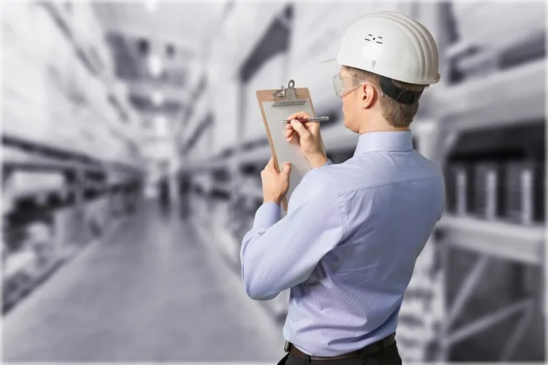 A person wearing a white hard hat and safety glasses stands in a warehouse, holding a clipboard and pen, appearing to take notes or conduct an inspection—perhaps documenting conditions for a personal injury attorney. The background shelves are blurred.