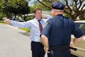 A man in a shirt and tie stands on a residential street with his arms outstretched, touching his nose, while a police officer observes—sobriety tests like this can help prevent car accidents and may require the guidance of lawyers.