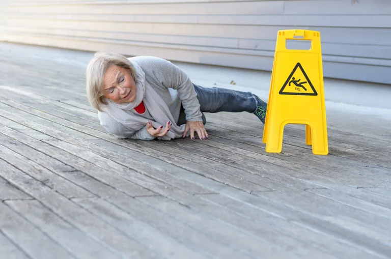 An older woman lies on a wooden floor after a fall, looking distressed. A yellow caution sign with a slipping person icon is nearby, highlighting the risk of personal injury from slippery surfaces.