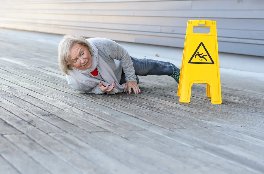 An older woman lies on a wooden floor after a fall, looking distressed. A yellow caution sign with a slipping person icon is nearby, highlighting the risk of personal injury from slippery surfaces.