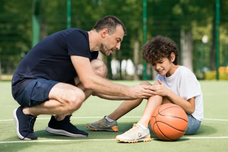 A man kneels beside a boy on an outdoor basketball court, gently examining the boy’s injured knee—much like lawyers or a car accident attorney would carefully assess injuries after car accidents. A basketball rests near the boy.