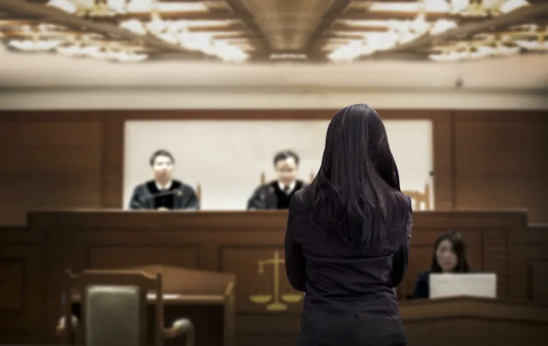 A woman with long dark hair stands facing three judges seated at a bench in a courtroom, while a personal injury attorney sits nearby, writing at a desk.