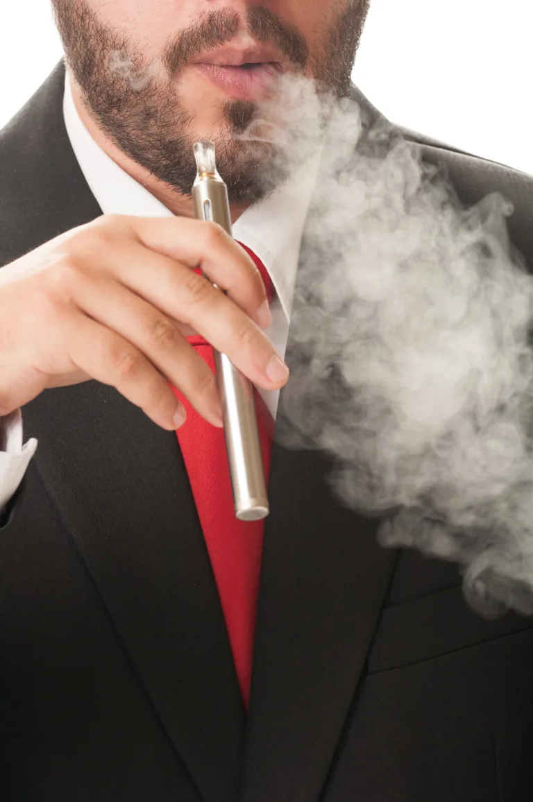 A man in a suit and red tie exhales vapor while holding an electronic cigarette close to his mouth, thick clouds partially obscuring his face—much like a personal injury attorney navigating through challenging legal battles.