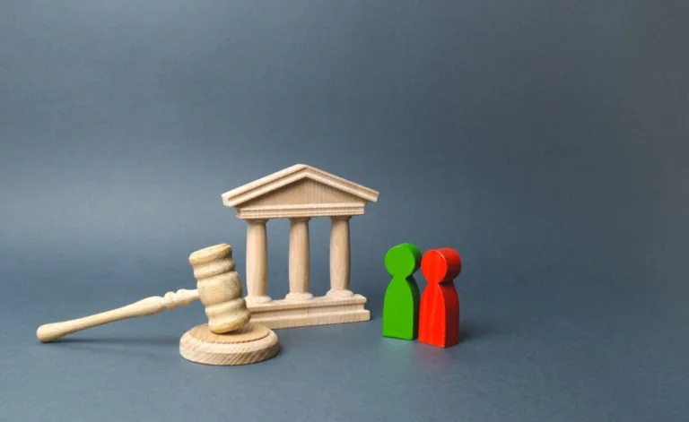 Wooden figures of a man and woman stand near a gavel and courthouse model, symbolizing a legal proceeding like divorce or dispute resolution, where lawyers or a personal injury attorney may offer guidance. The background is plain gray.