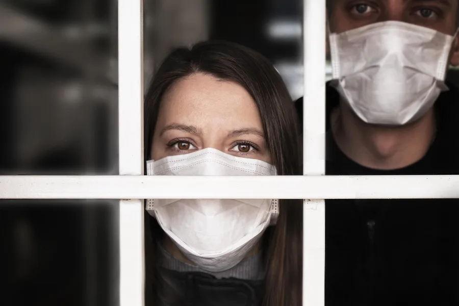 Two people wearing white face masks look out from behind a window with white bars. The woman gazes ahead, while the man stands behind her. The scene evokes isolation—much like the aftermath of car accidents or seeking help from a personal injury attorney.