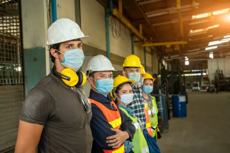 A group of factory workers wearing hard hats, safety vests, and face masks stand side by side inside an industrial warehouse, looking in the same direction—much like a team of personal injury attorneys ready to advocate for workplace safety.