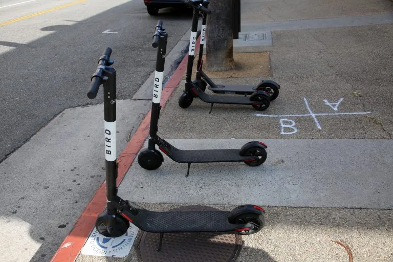 Three black Bird electric scooters are parked on a sidewalk near a curb. White chalk markings form a tic-tac-toe grid with a “B” and “Y” on the pavement—reminding passersby of safety concerns often addressed by personal injury lawyers.