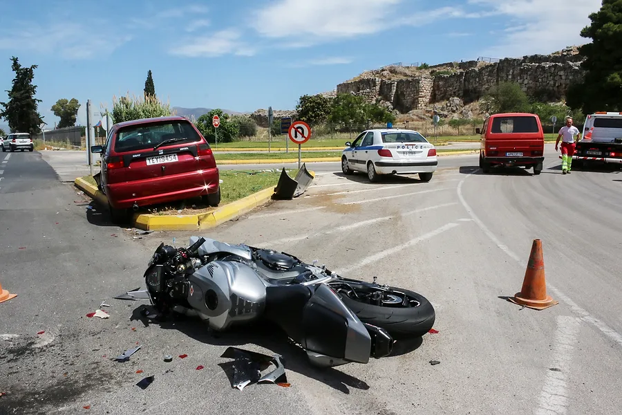A silver motorcycle lies on its side in the road after an accident near a red car, which is up on a curb. Police cars and officers are present, and traffic cones block part of the scene as lawyers or a car accident attorney may be needed.