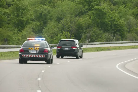 A police car with flashing lights follows a black SUV on a curving road surrounded by green trees, highlighting moments when personal injury lawyers are needed most.